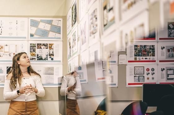 Photo of a Chatham University female student looking up at printed out design projects hung on the wall. 