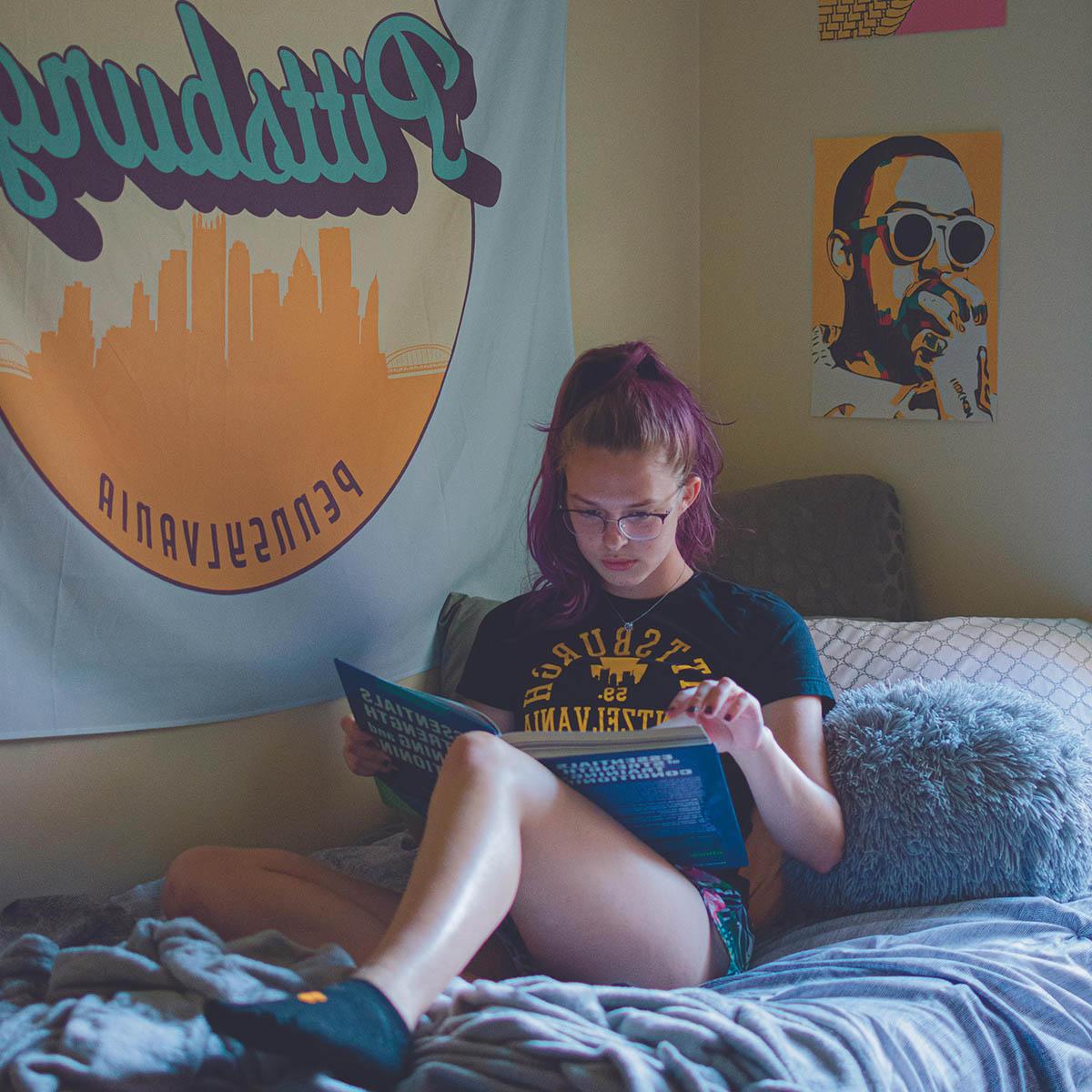 照片中，一位年轻的女学生梳着粉红色的马尾辫，戴着眼镜，躺在宿舍的床上看书. 墙上挂着一幅写着宾夕法尼亚州匹兹堡市的大挂毯