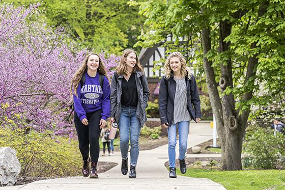 春季，波胆网站的三名女学生在足球波胆平台步行去上课, 有绿色的树和五颜六色的花. 